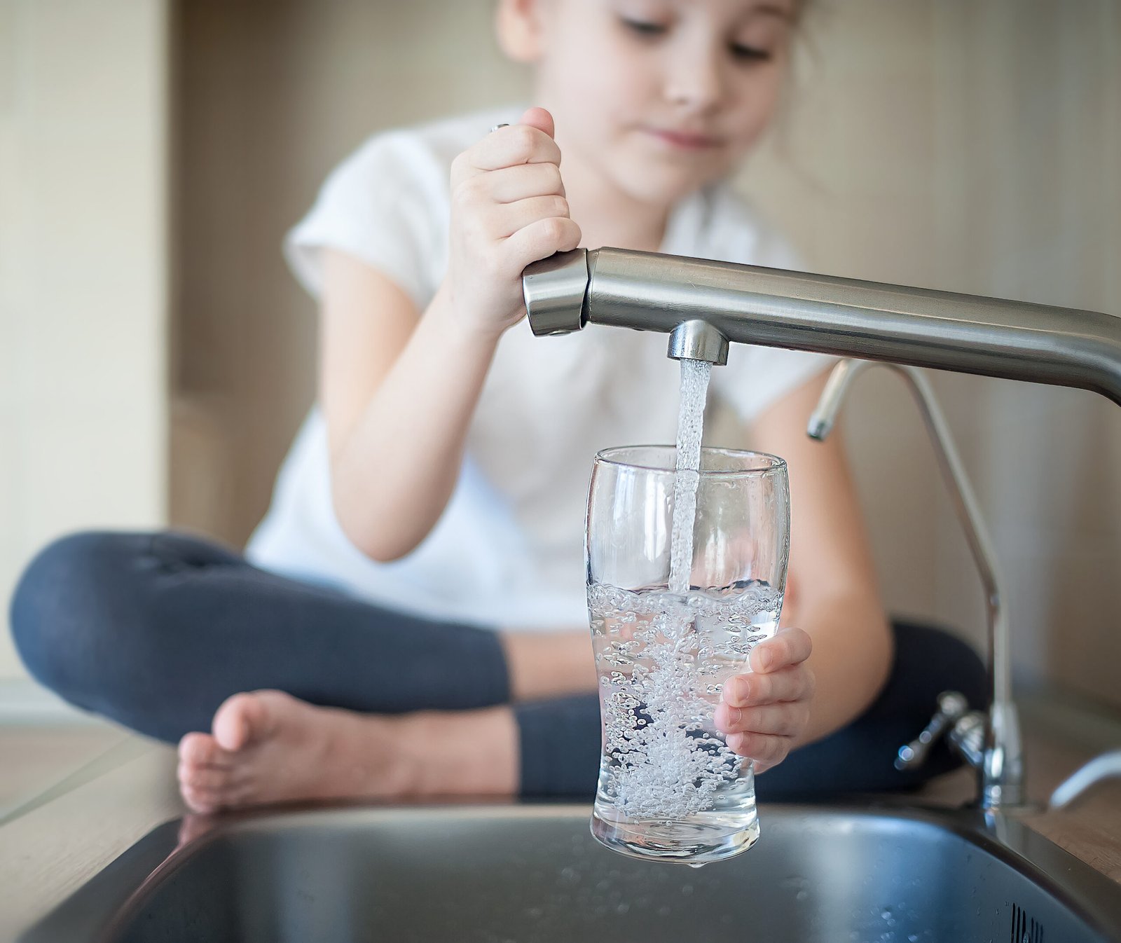 Little girl opens a water tap with her hand holding a transparent glass. Kitchen faucet. Glass of clean water. Filling cup beverage. Pouring fresh drink. Hydration. Healthcare. Healthy lifestyle.